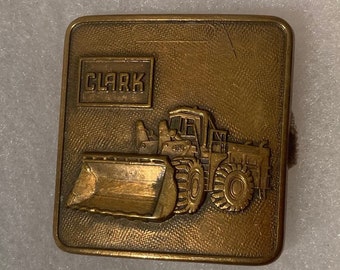 Puede incluir: Una hebilla de cinturón de metal dorado con una imagen detallada de una cargadora frontal Clark con un logotipo "CLARK" en relieve.