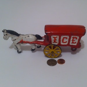 May include: A vintage cast iron toy ice cream wagon pulled by a white and black horse. The wagon is red with white lettering that says "ICE".