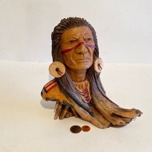 May include: A wooden sculpture of a Native American man's head with red and white paint on his face. The sculpture is on a wooden base and has a beaded necklace.