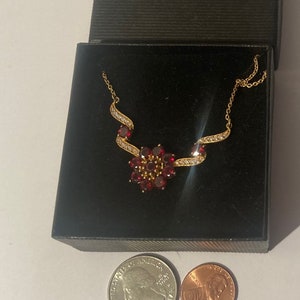 May include: A gold necklace with a red gemstone pendant in a black velvet box. The pendant is a cluster of red gemstones in a flower shape.