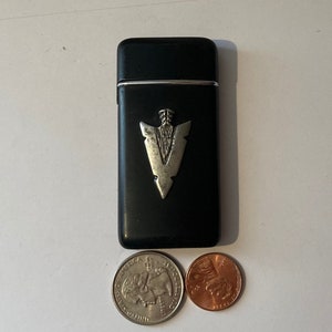May include: A black leather cigarette lighter with a silver arrowhead design. The lighter is about the size of a US quarter.