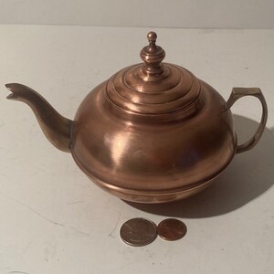 Könnte beinhalten: Eine Kupferteekanne mit rundem Korpus und einem gewölbten Deckel. Die Teekanne hat einen Ausguss und einen Griff. Die Teekanne steht auf einer weißen Oberfläche.