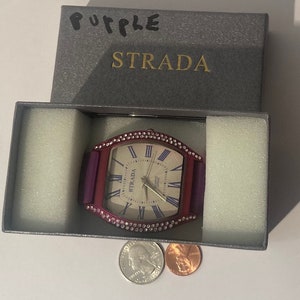 May include: A purple watch with a white face and silver hands. The watch is in a gray box with the word "STRADA" printed on it.