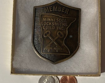 May include: A bronze-coloured metal badge with the text "MEMBER MINNESOTA LOCKSMITHS GUILD INC." and a stylised image of the state of Minnesota with two crossed keys. The badge is in a white box with a gold trim.