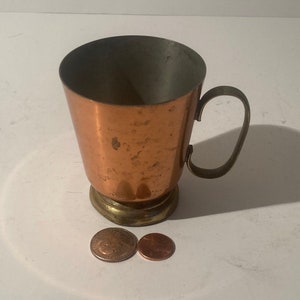 Puede incluir: Una taza de cobre con asa y base de latón. La taza tiene un acabado martillado y está sobre una superficie blanca con dos monedas de cobre.