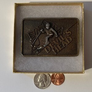 May include: A bronze belt buckle with a skier in a silver finish and the words "Devil's Head" in gold lettering.