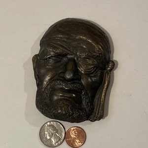 May include: A bronze sculpture of a man's face with a bandage over one eye. The sculpture is detailed and shows the man's facial features, including his beard and mustache.