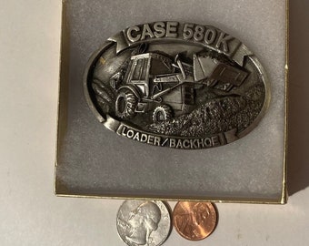 Puede incluir: Hebilla de cinturón de metal plateado con un Case 580K Loader/Backhoe. La hebilla es de forma ovalada y está colocada en una pequeña caja con borde dorado. Un cuarto de dólar y un centavo están al lado.