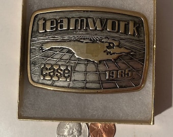 Puede incluir: Una hebilla de cinturón de oro y plata con las palabras "teamwork" y "case" y el año "1965" en ella. La hebilla presenta un mapa de un estado con un patrón de cuadrícula.