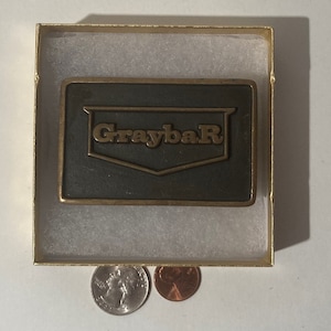 May include: A rectangular metal belt buckle with the word "GraybaR" in gold lettering. The buckle is dark green and set in a gold-colored box with a white interior. Two coins are visible below the box.