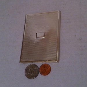 May include: A silver compact with a ridged surface and a rectangular indentation. The compact is about the size of two US coins.