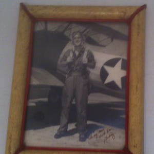 May include: A black and white photo of a man in a military uniform standing in front of a vintage airplane. The photo is framed in a wooden frame with a red and gold border. The man is smiling and holding a hat in his hand. The photo is signed "In my next life, I'm a pilot."