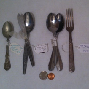 Puede incluir: Una colección de cubiertos vintage, que incluye una cuchara de plata, un juego de cucharas para servir y un tenedor. Los cubiertos se muestran sobre una superficie blanca con etiquetas de precio.