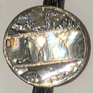 May include: Circular pendant with a silver-colored metal frame. The pendant's center features iridescent abalone shell segments, displaying a range of colors. The pendant is attached to a black cord, likely for wearing as a necklace or bolo tie.