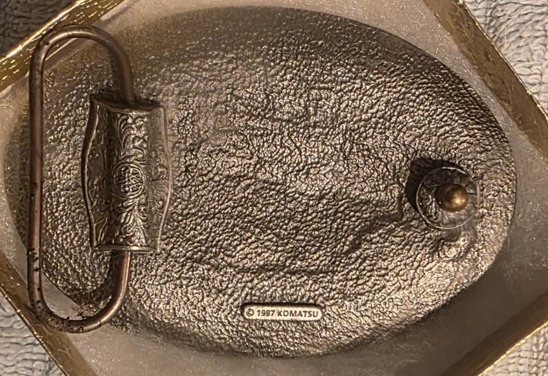 Op de afbeelding: Een vintage metalen riemgesp met een getextureerd, ovaal ontwerp. De gesp heeft een sierlijke, decoratieve sluiting en een kleine, ronde knop. De tekst "&copy; 1997 KOMATSU" is zichtbaar op de achterkant. De gesp zit in een goudkleurige doos.
