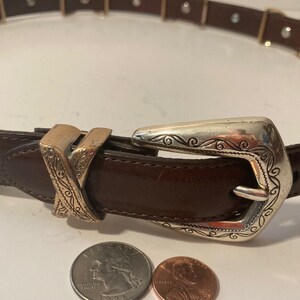 Puede incluir: Un cintur&oacute;n de cuero marr&oacute;n con una hebilla plateada y dorada. La hebilla tiene un dise&ntilde;o adornado con espirales y l&iacute;neas. El cintur&oacute;n est&aacute; cerrado y la hebilla est&aacute; abrochada.