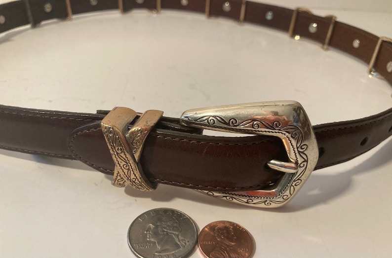 Puede incluir: Un cintur&oacute;n de cuero marr&oacute;n con una hebilla plateada y dorada. La hebilla tiene un dise&ntilde;o adornado con espirales y l&iacute;neas. El cintur&oacute;n est&aacute; cerrado y la hebilla est&aacute; abrochada.