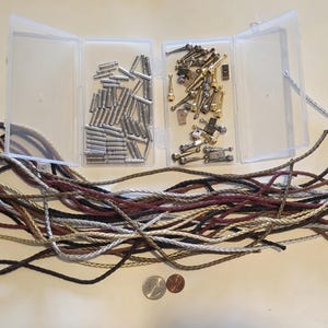 Op de afbeelding: Een assortiment benodigdheden voor het maken van sieraden, waaronder zilver- en goudkleurige metalen fournituren in doorzichtige plastic containers. Gevlochten koorden in verschillende kleuren, waaronder zwart, bruin en bordeaux, zijn ook aanwezig. Twee munten zijn zichtbaar aan de onderkant.