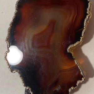May include: A brown agate stone bolo tie with a gold-toned metal trim. The stone has a natural, irregular shape and features a swirling pattern of brown, white, and orange.