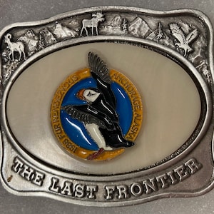 Puede incluir: Hebilla de cinturón de plata con un fondo ovalado blanco que presenta un frailecillo colorido con el texto "1988 Fur Rendezvous Anchorage Alaska". La hebilla también tiene el texto "The Last Frontier" en la parte inferior.