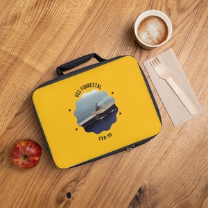 Könnte beinhalten: Gelbe Lunchbox mit schwarzem Reißverschluss und schwarzem Griff. Die Tasche zeigt eine Grafik des Flugzeugträgers USS Forrestal mit dem Text "USS FORRESTAL CVA-59".