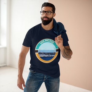 Może przedstawiać: Granatowa koszulka z okrągłą grafiką z napisem "USS MCKEE" i przedstawieniem statku na morzu. Grafika wykorzystuje odcienie niebieskiego, turkusowego, pomarańczowego i czarnego. Koszulka wykonana jest z miękkiego materiału.