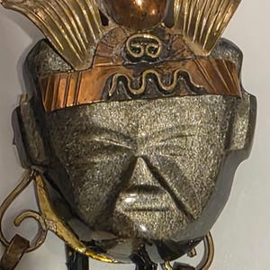 Op de afbeelding: Een gedetailleerde close-up van een metalen sculptuur die een gestileerd gezicht met een hoofdtooi afbeeldt. De sculptuur heeft goud-, koper- en zilvertinten. Het gezicht heeft duidelijke kenmerken en een serene uitdrukking. Het stuk lijkt een artistieke representatie te zijn.