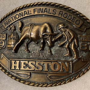 May include: A bronze belt buckle with a rope border. The buckle features a scene of a cowboy roping a bull. The buckle also has the text "National Finals Rodeo" and "NFR 19 81" and the name "Hesston".