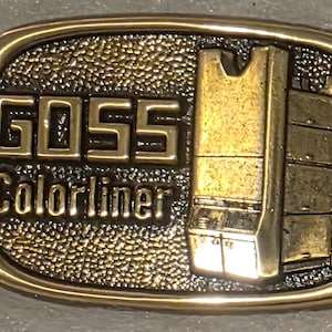 Könnte beinhalten: Eine Vintage-Gürtelschnalle aus Messing mit dem Text "GOSS Colorliner" und einer detaillierten Abbildung eines Fahrzeugs. Die Schnalle hat einen strukturierten Hintergrund und einen goldfarbenen Rand, präsentiert in einer Box.