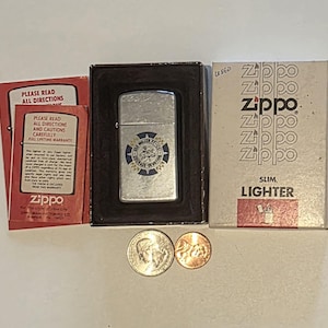 以下が含まれることがあります： 青と白のエンブレムが付いたシルバーのZippoライター。黒い箱に入っており、「SLIM LIGHTER」と書かれた箱の隣にあります。25セント硬貨と1セント硬貨も含まれています。「すべての指示をお読みください」というテキストの赤いパンフレットが見えます。