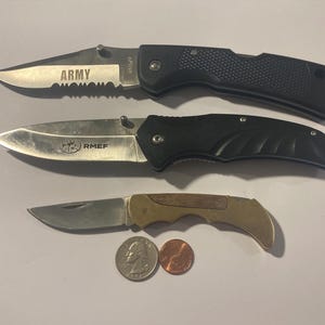 May include: Three folding knives with silver blades and black or brown handles are displayed. The top knife has the word "ARMY" etched on the blade. The middle knife has the letters "RMEF" etched on the blade. Two coins are next to the bottom knife.