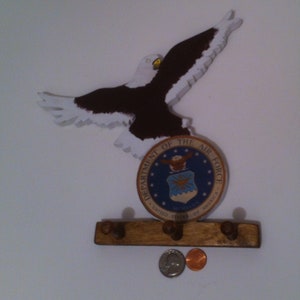 Könnte beinhalten: Ein hölzerner Adler-Wandhaken mit einem braun-weiß bemalten Adler-Design. Der Adler fliegt mit ausgebreiteten Flügeln. Der Haken hat eine braune Holzbasis mit vier Haken. Ein rundes Siegel mit dem Text "Department of the Air Force" und dem Emblem der US Air Force ist an der Basis befestigt.