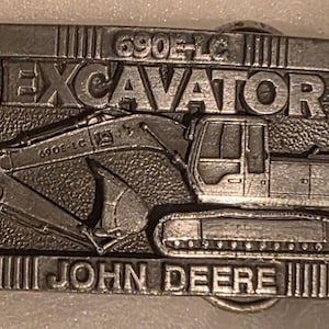 May include: A rectangular, silver-colored belt buckle featuring an excavator design. The word "EXCAVATOR" is prominently displayed above the excavator image, with "JOHN DEERE" below. The model number "690E-LC" is also visible.