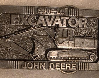 Puede incluir: Hebilla de cinturón rectangular plateada con un diseño de excavadora. La palabra "EXCAVATOR" se muestra prominentemente sobre la imagen de la excavadora, con "JOHN DEERE" debajo. También es visible el número de modelo "690E-LC".