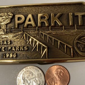 Może przedstawiać: Brązowa klamra do paska w kształcie stanu Kansas z napisem "PARK IT" i "KANSAS STATE PARKS 1986". Klamra zawiera również mały obrazek drzewa i schodów.
