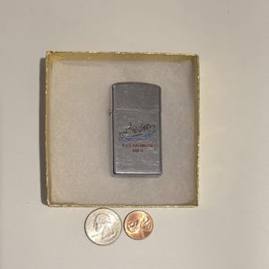 May include: A silver-colored lighter with a ship design and the text "U.S.S. Kalamazoo AOR-6" in blue and red. The lighter is in a gold-colored box, with a quarter and a penny next to it.