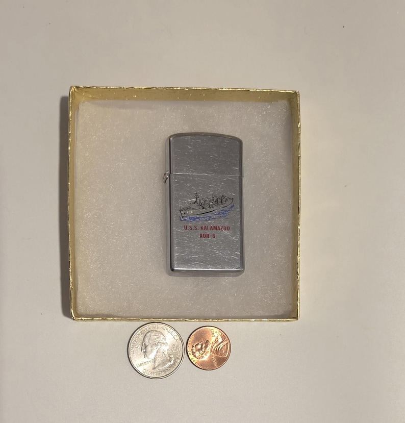 May include: A silver-colored lighter with a ship design and the text "U.S.S. Kalamazoo AOR-6" in blue and red. The lighter is in a gold-colored box, with a quarter and a penny next to it.