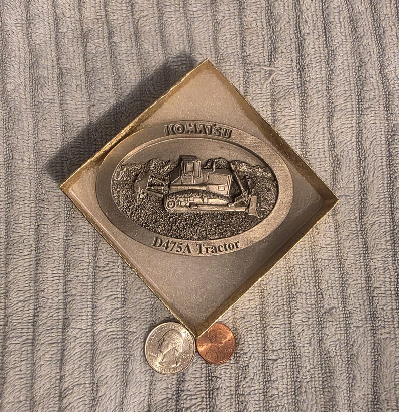 Op de afbeelding: Een zilverkleurig Komatsu D475A Tractor embleem in een goudkleurige doos. Het embleem toont een gedetailleerd tractorontwerp. Een kwart dollar en een cent worden naast de doos geplaatst ter vergelijking.