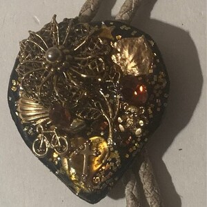 Könnte beinhalten: Ein herzförmiger Bolo-Schlips mit einem gold-schwarzen Design. Das Design zeigt eine Vielzahl von kleinen Objekten, darunter ein Fahrrad, einen Vogelkäfig und einen kleinen Baum. Der Bolo-Schlips ist aus Leder und hat eine geflochtene Schnur.