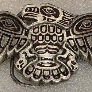 May include: A silver-colored metal belt buckle featuring an eagle design with intricate details. The eagle has outstretched wings and a detailed head, with dark brown accents. The buckle is a rectangular shape with a curved bottom.