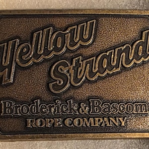 May include: A rectangular, bronze-colored belt buckle with the words "Yellow Strand" in a stylized font. Below, it reads "Broderick & Bascom Rope Company". The buckle has a textured surface and a vintage aesthetic.