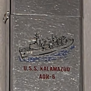 May include: A silver-colored lighter with a ship illustration and the text "U.S.S. Kalamazoo AOR-6". The ship is depicted in black and blue, with the text in red. The lighter has a shiny, reflective surface.