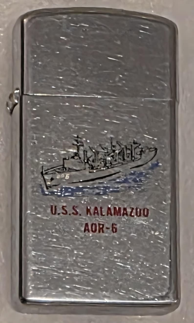 May include: A silver-colored lighter with a ship illustration and the text "U.S.S. Kalamazoo AOR-6". The ship is depicted in black and blue, with the text in red. The lighter has a shiny, reflective surface.