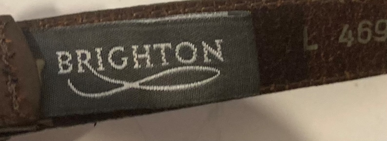 Puede incluir: Un cintur&oacute;n de cuero marr&oacute;n con una etiqueta gris que dice "BRIGHTON" y una etiqueta blanca con el n&uacute;mero "L 4699".