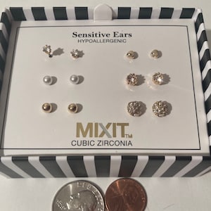 Könnte beinhalten: Eine weiße Schachtel mit schwarz-weißen Streifen enthält ein Set mit 10 goldenen und perlmuttartigen Ohrringen. Die Ohrringe sind hypoallergen und mit Cubic Zirconia Steinen besetzt. Die Schachtel trägt den Text "Sensitive Ears Hypoallergenic" und "Mixit™ Cubic Zirconia".