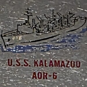 May include: A stylized illustration of the U.S.S. Kalamazoo AOR-6 naval vessel. The image features a detailed depiction of the ship, with the ship's name and designation in red text below the ship.