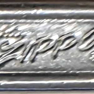 May include: Close-up of a silver metal lighter base with the embossed brand name "Zippo" in a cursive font. The lighter base has a rectangular shape with rounded edges and a textured surface.