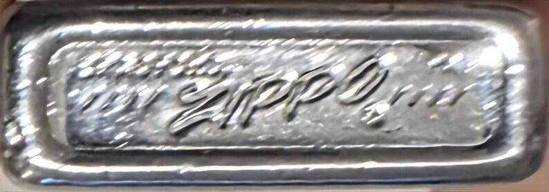 May include: Close-up of a silver metal lighter base with the embossed brand name "Zippo" in a cursive font. The lighter base has a rectangular shape with rounded edges and a textured surface.
