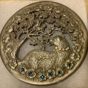 Puede incluir: Una pieza decorativa circular plateada con un relieve detallado de un perro descansando bajo un árbol en flor. El collar del perro está adornado con pequeñas gemas iridiscentes, y las flores están acentuadas con gemas azules.