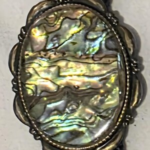 Könnte beinhalten: Ovaler Bolo-Krawattenanhänger mit einer schillernden Abalone-Muscheleinlage. Die Muschel zeigt eine Reihe von Farben, darunter Gold, Grün und Blau, eingebettet in einen dekorativen silberfarbenen Rahmen. Die Bolo-Krawatte hat eine dunkelblaue geflochtene Kordel.